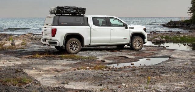 A white Sierra truck equipped with accessories parked on a rocky beach next to a body of water